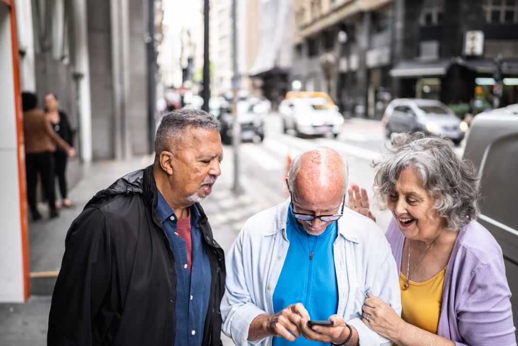 Drie oudere mensen die samen lachend naar een smartphone kijken en deze gebruiken op een stadsstoep