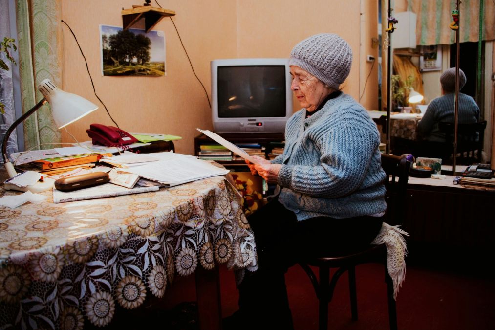 Oudere persoon zittend aan een tafel die een document leest in een verlichte kamer met een tv op de achtergrond en wanddecoraties