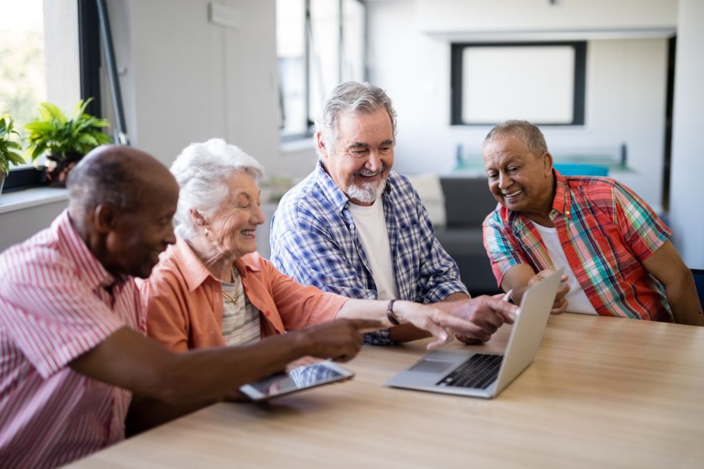Vier glimlachende oudere mensen zittend rond een tafel en samen kijkend naar een laptop